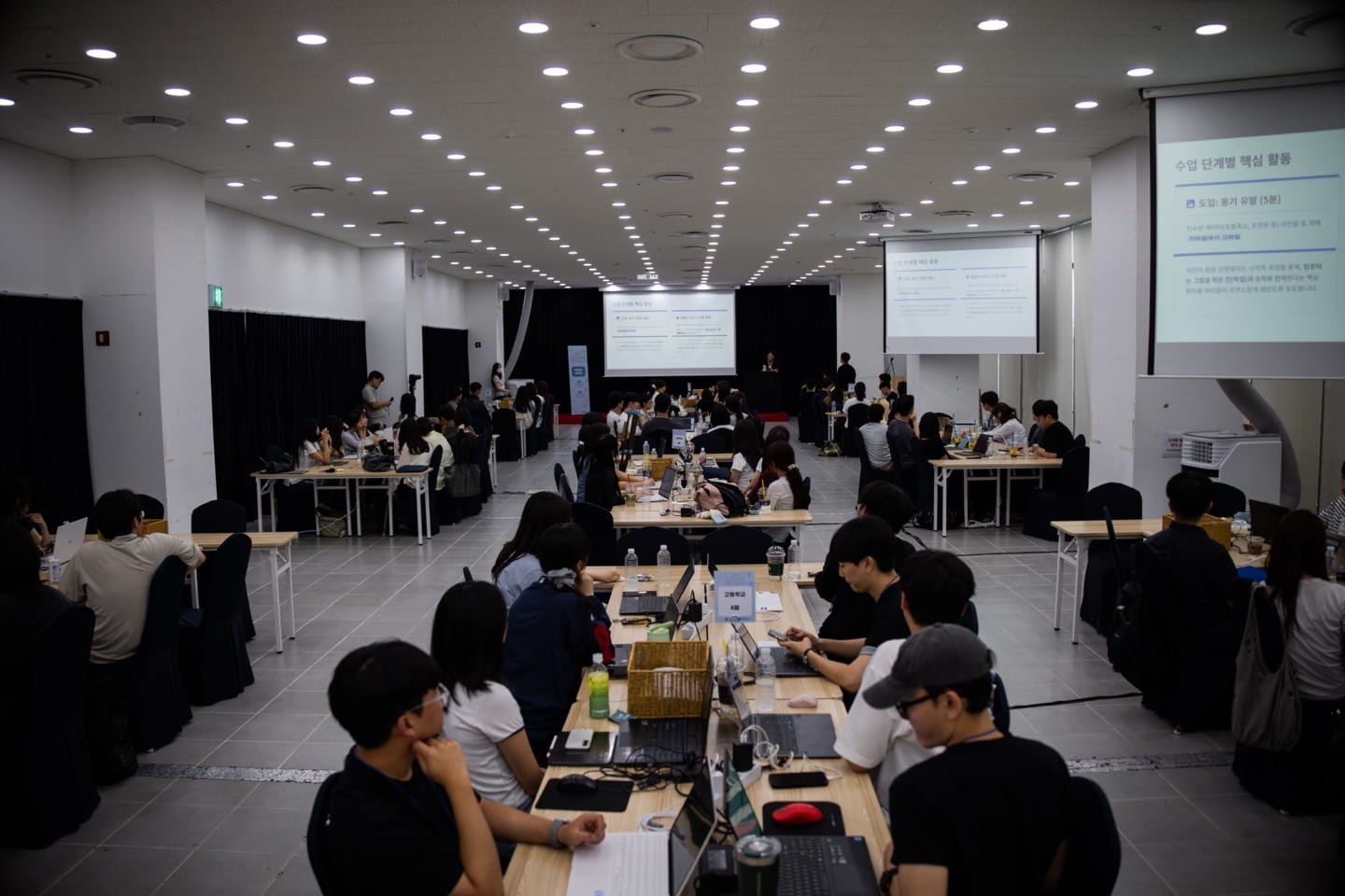 워크숍장 전경 — 팀별로 나뉜 긴 테이블에서 멘토들이 노트북과 활동지를 놓고 작업하고 있다